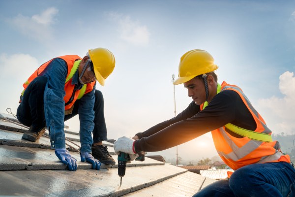 Construction worker wearing safety harness belt during working on roof structure of building on construction site,Roofer using air or pneumatic nail gun and installing concrete roof tile on top roof.