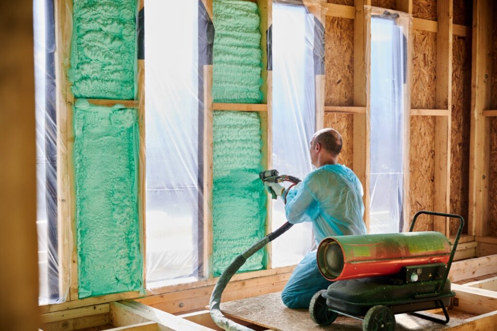 Male builder insulating wooden frame house. Man worker spraying polyurethane foam inside of future cottage, using plural component gun