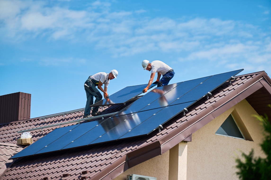 Workers building solar panel system on rooftop of house. Two men installers in helmets installing photovoltaic solar module outdoors. Alternative, green and renewable energy generation concept.