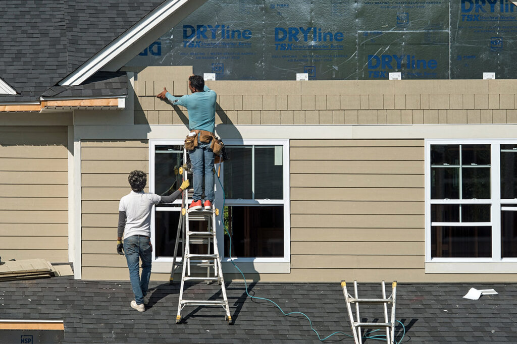 Two men installing siding on a house using a ladder, demonstrating seasonal home improvement techniques.